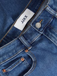 Jeans de mezclilla en azul medio, con una etiqueta que dice "JJXX", remaches de cobre, costuras de color marrón dorado y un diseño clásico de cinco bolsillos.