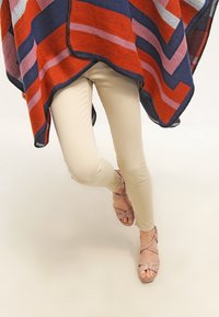 Osoba oblečena v béžové úzké kalhoty, růžové sandály na klínku se š straps, a pončo s pruhy v červené, námořnické modré a růžové barvě.