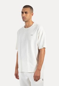 Biała, oversize koszulka z bawełny, z okrągłym dekoltem, krótkimi rękawami i małą metką z logo na piersi. Gładka tekstura.