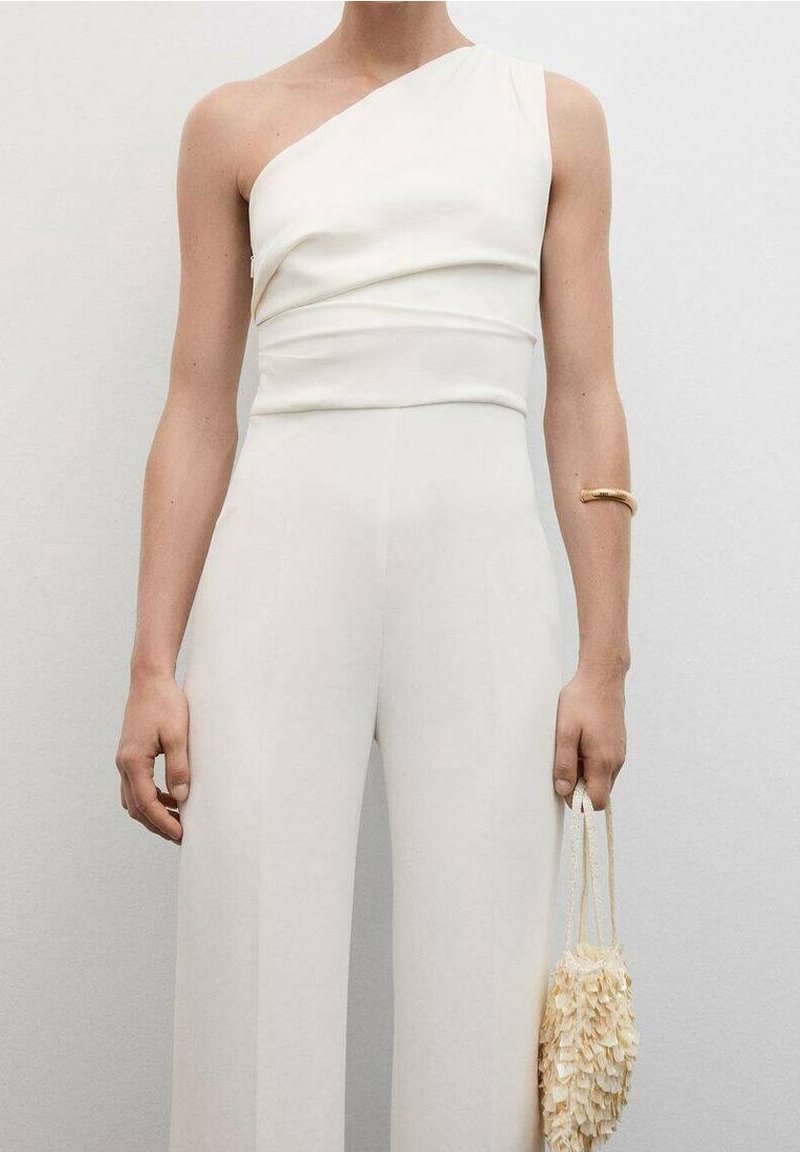 Personne portant une combinaison blanche asymétrique et un bracelet en or, tenant un petit sac à main beige texturé à côté d'elle.