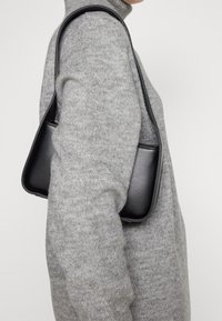 Borsa a tracolla in pelle nera con una silhouette triangolare e una tracolla sottile, indossata sopra un maglione grigio chiaro in maglia testurizzata.