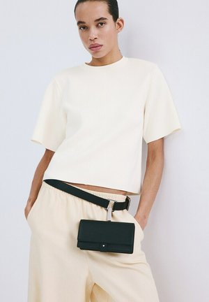 Mujer con camiseta de manga corta color crema y pantalones con cinturón negro tipo riñonera, apoyada contra una pared blanca con las manos en los bolsillos.