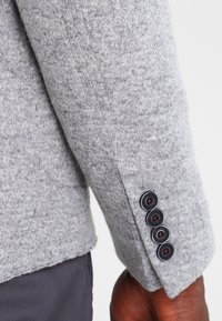 Manga de blazer gris texturizado con cuatro botones negros en un brazo de piel oscura, combinado con pantalones gris oscuro.