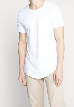 Mann trägt ein schlichtes weißes T-Shirt mit kurzen Ärmeln und Brusttasche sowie hellbeige Hose vor einfachem hellem Hintergrund.