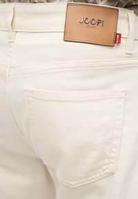 Κοντινό πλάνο πίσω τσέπης κρεμ τζιν με ταμπά δερμάτινη ετικέτα JOOP! Jeans ραμμένη από πάνω.