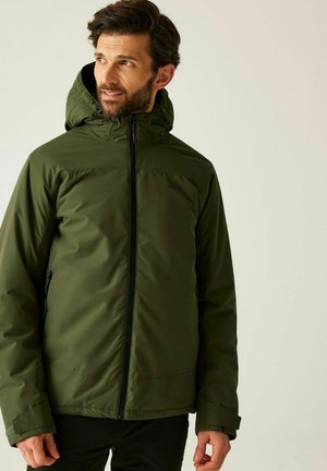 Hombre con chaqueta verde con capucha y cremallera, de pie y mirando ligeramente hacia la izquierda sobre un fondo claro y sencillo.