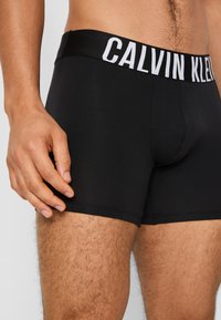 Crne bokserice od glatke tkanine, s bijelim pojasom na kojem piše "CALVIN KLEIN", uskog kroja, kratkih nogavica.