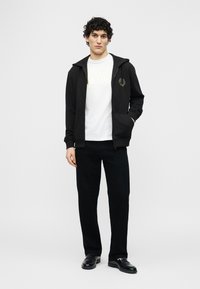 Νέος άνδρας με σγουρά μαλλιά, φορώντας λευκό μπλουζάκι, μαύρο φούτερ με φερμουάρ, μαύρο παντελόνι και μαύρα παπούτσια, στέκεται με το ένα χέρι στην τσέπη.