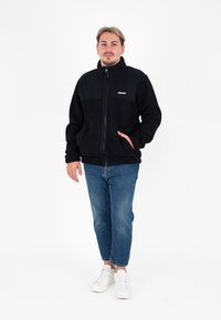 Giacca in pile nera con zip, caratterizzata da una parte superiore nera uniforme e un logo. Abbinata a jeans azzurri chiari e sneakers bianche.
