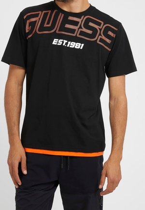 Homme portant un T-shirt noir Guess avec un logo orange en contour contenant le texte "GUESS" et "EST.1981" en blanc, assorti à un pantalon noir.