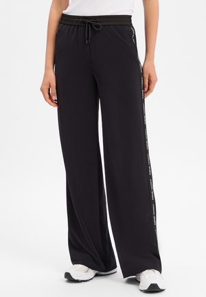 Pantalon noir à jambes larges avec taille à cordon et baskets blanches, personne debout devant un fond uni.