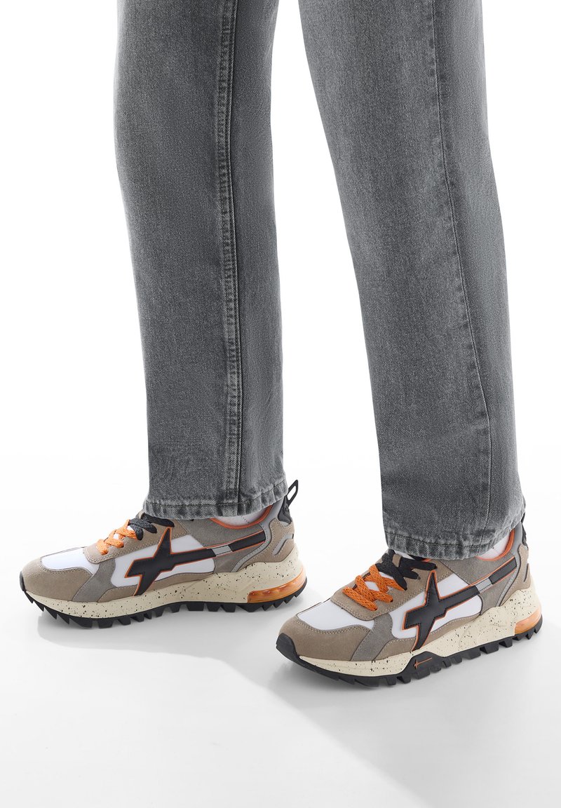 Jeans grigi indossati con sneakers beige, bianche e nere con dettagli e lacci arancioni, in piedi su una superficie bianca.
