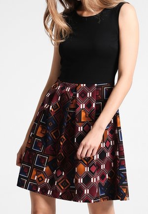 Kvinde iført sort ærmeløs top og knælange nederdel med geometriske mønstre i bordeaux, orange, marineblå og hvid.