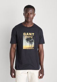 Camiseta de algodón negra con un estampado gráfico de palmeras y un atardecer, con el texto "GANT Resort" destacado.