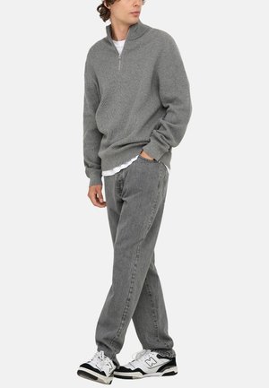 Grauer, gerippter Pullover mit einem Halbzip-Kragen, getragen über einem weißen Hemd, kombiniert mit faded grauen Jeans und schwarz-weißen Turnschuhen.