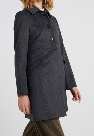 Femme portant un manteau en laine gris foncé boutonné avec des poches latérales et un col, debout devant un fond clair uni.