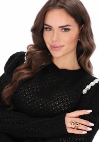 Fekete kötött pulóver texturált, nyitott szövésmintával és gyöngydíszítéssel a vállakon. Ujjain sima bőrszínű gyűrűk, természetes körmök.