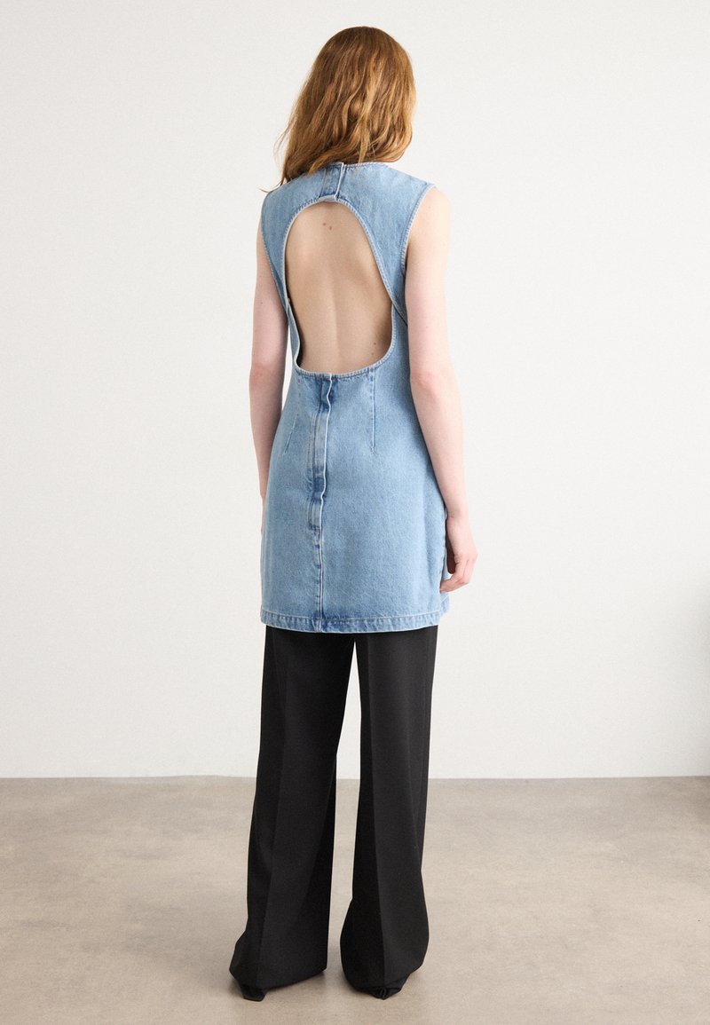 Τζιν φόρεμα με ανοιχτή πλάτη, αμάνικο σχέδιο και ανοιχτό μπλε χρώμα. Συνδυασμένο με φαρδύ μαύρο παντελόνι, αναδεικνύει μια χαλαρή σιλουέτα.