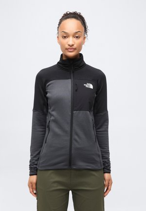 Γυναίκα με σγουρά μαλλιά που φοράει μαύρο και γκρι φλις μπουφάν με φερμουάρ και ελαφρώς χακί παντελόνι, στέκεται μπροστά σε ανοιχτό γκρι φόντο.