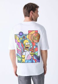 Hvid kortærmet t-shirt med et farverigt grafik af en tegneseriefigur, der holder en drink, omgivet af abstrakte former og levende mønstre.
