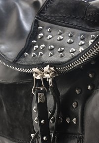 Sac en cuir noir avec des clous en métal argenté, une fermeture éclair et une surface texturée. Les accessoires comprennent une double fermeture éclair et une étiquette de marque.