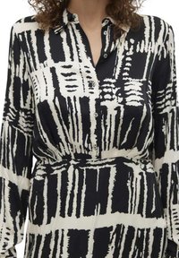 Robe chemise à motifs noir et blanc avec un col, fermeture à boutons, manches longues et détail de taille froncée. Texture de tissu lisse.