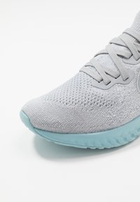 Šedá pletená sportovní bota s texturovaným svrškem, plochými tkaničkami a světlemodrou gumovou podrážkou s vlnitým vzorem.