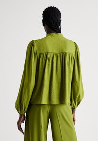 Blusa verde con texture, maniche a sbuffo e dettagli arricciati sul retro; abbinata a pantaloni larghi verdi coordinati.