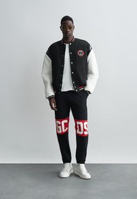 Black and white varsity jacket with red accents, paired with black pants featuring large red and white lettering. Model wears white sneakers.