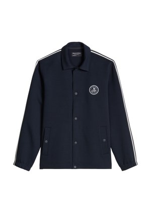 Mann in navyblauer Jacke und Hose mit weißem Logo, beige Kappe mit schwarzem Schirm, weißem Hemd und weißen Sneakers steht mit den Händen in den Taschen.