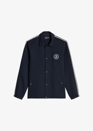 Bărbat în jachetă și pantaloni bleumarin cu logo alb, șapcă bej cu vizieră neagră, cămașă albă și adidași albi, stând cu mâinile în buzunare.