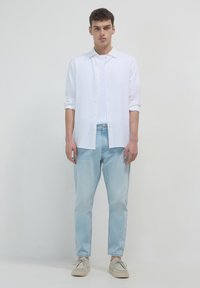 Giovane uomo che indossa una camicia bianca con bottoni sopra una maglietta bianca, jeans azzurri chiari e scarpe casual beige, in piedi di fronte a uno sfondo neutro.