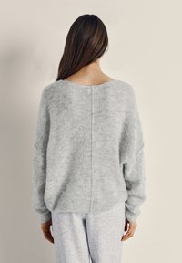 Žena s dlhými hnedými vlasmi, oblečená v voľnom svetlosivom pletenom svetri a svetlosivých nohaviciach, stojaca čelom k jednoliatej béžovej stene.