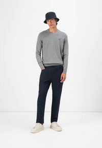 Giovane uomo che indossa un maglione grigio a V con logo giallo, pantaloni blu navy, sneakers bianche e un cappello a secchiello nero, in piedi su uno sfondo bianco.