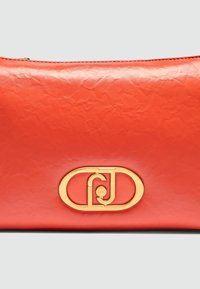 Rød læder clutch med en struktureret finish, prydet med et guldfarvet oval logo i midten. Lynlås lukning øverst.