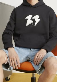 Felpa nera con un design a forma di fulmine bianco e logo Jordan, indossata con pantaloni shorts in denim blu sfrangiati, seduto su una sedia arancione.