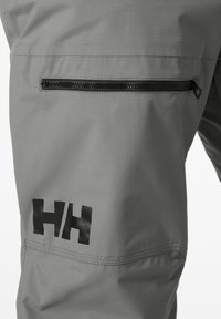 Grijze waterdichte broek heeft een ritszak aan de zijkant, gestructureerde stof en een opvallend zwart logo op de bovenbeen. Duurzaam en functioneel ontwerp.