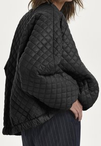 Person i en oversized sort quiltet jakke med knaplukning ved manchetterne og mørke stribede bukser foran en ensfarvet lys baggrund.