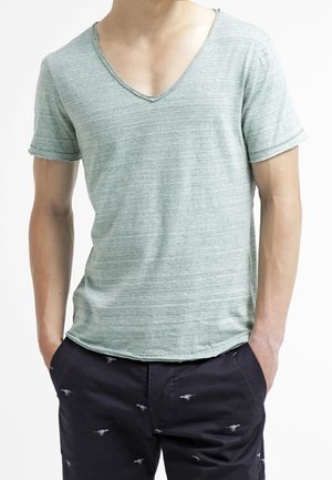 Mężczyzna w jasnoszarym T-shircie z dekoltem w serek i krótkim rękawem oraz ciemnych szortach z małymi zwierzęcymi wzorami, z rękami w kieszeniach, pokazany od szyi do bioder.