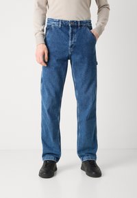 Jeans de mezclilla azul con un diseño de pierna recta, costuras visibles y bolsillos laterales. Combinados con zapatillas negras.