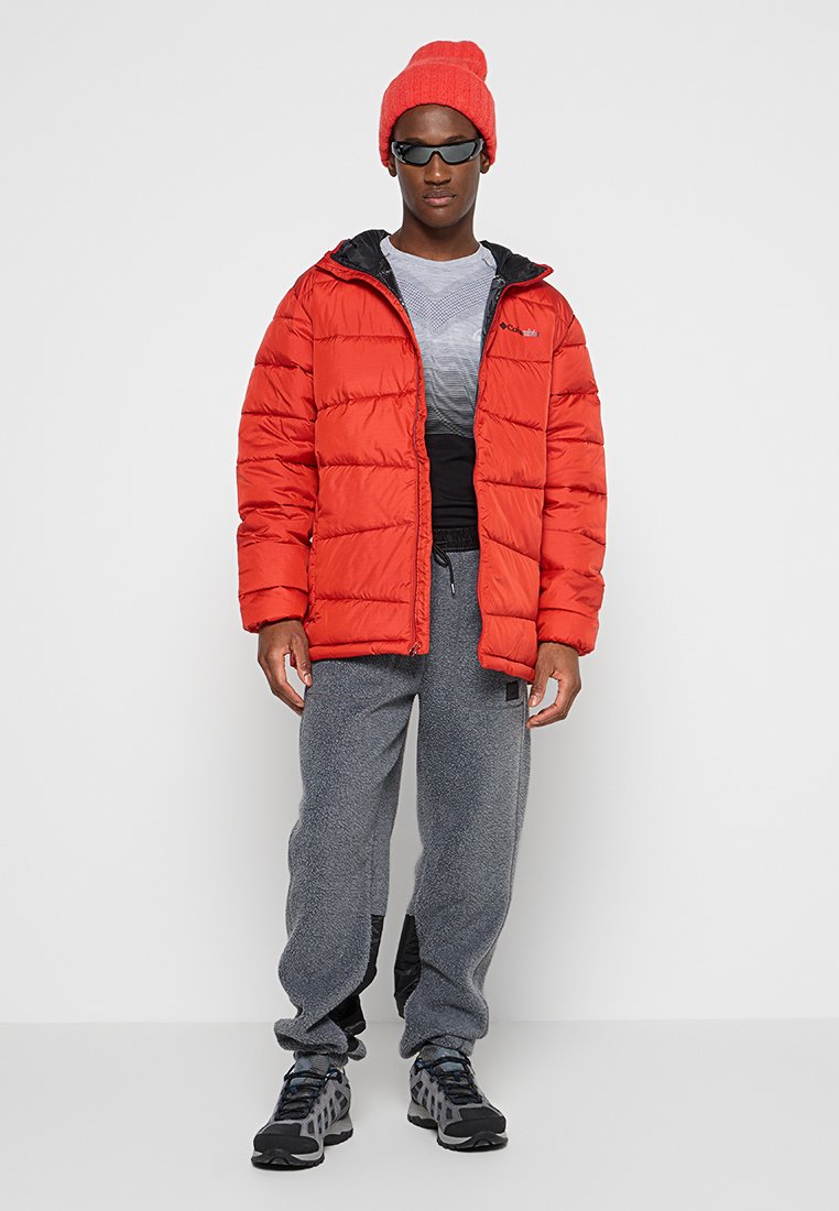 Chaqueta plumas roja con textura acolchada, forro negro, pantalones de forro polar grises y calzado negro. Los accesorios incluyen un gorro rojo y gafas de sol.