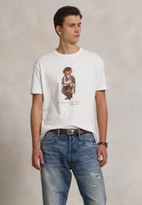 Biela bavlnená tričko s grafickým motívom medveďa v obleku. Nosené s obnošenými modrými džínsami a hnedým opaskom. Jednoduchý, neformálny dizajn.