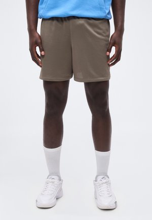 Persona con pantalones cortos deportivos de malla marrón, calcetines blancos y zapatillas blancas, de pie contra un fondo claro y liso.
