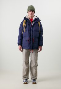 Veste en duvet marine avec des accents rouges, grandes poches et une capuche, associée à un pantalon cargo beige clair et un bonnet vert. Des baskets complètent le look.