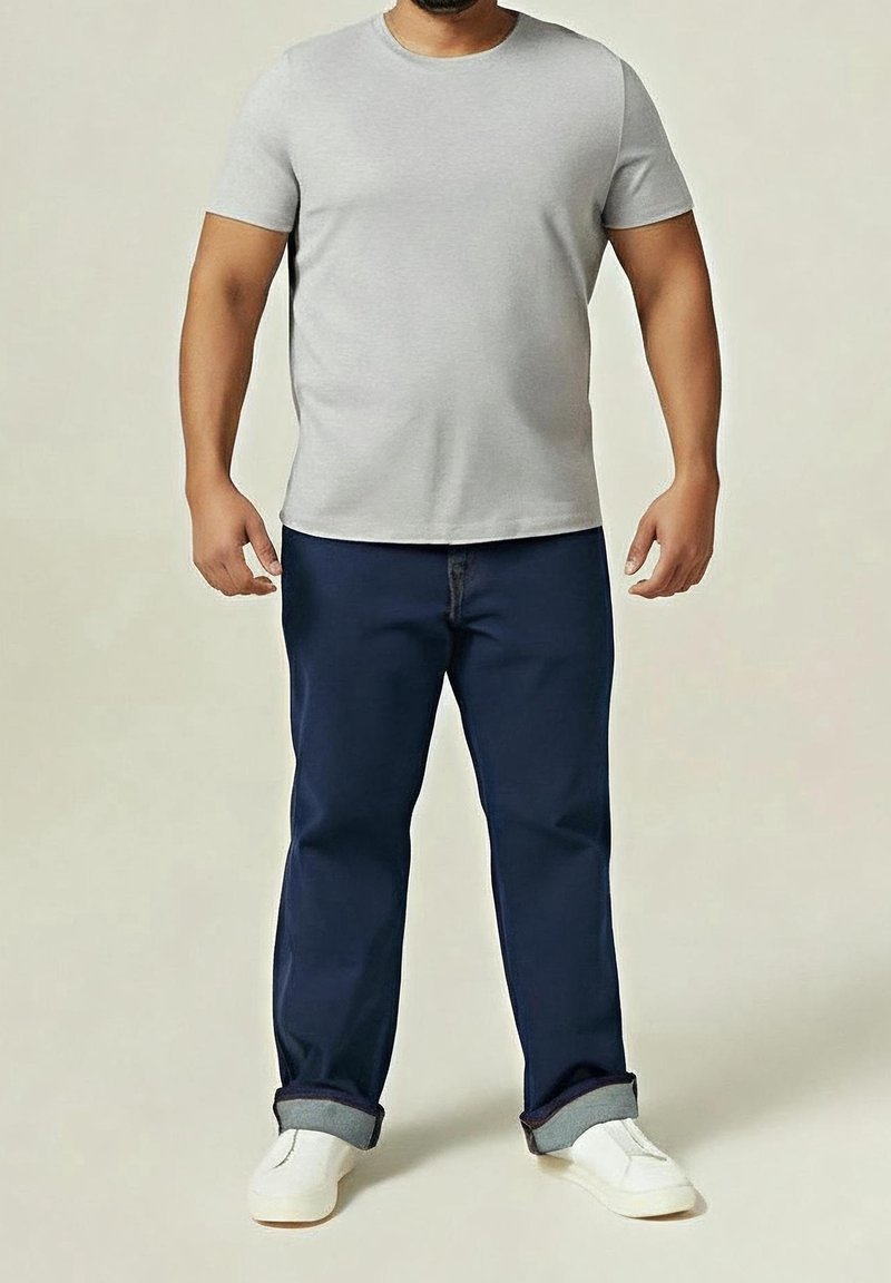 Homme portant un t-shirt gris clair à manches courtes, un jean bleu foncé retroussé et des baskets blanches, debout devant un fond uni.
