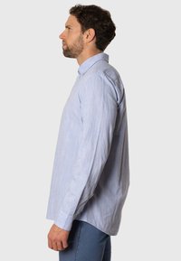 Camisa de manga larga azul clara hecha de una tela con una textura sutil. Presenta un cuello con botones y un corte holgado con aberturas laterales.