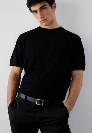 Mladi moški s kodrastimi lasmi, oblečen v preprosto črno kratko srajco, črne hlače in črni pas, stoji z rokami v žepih.