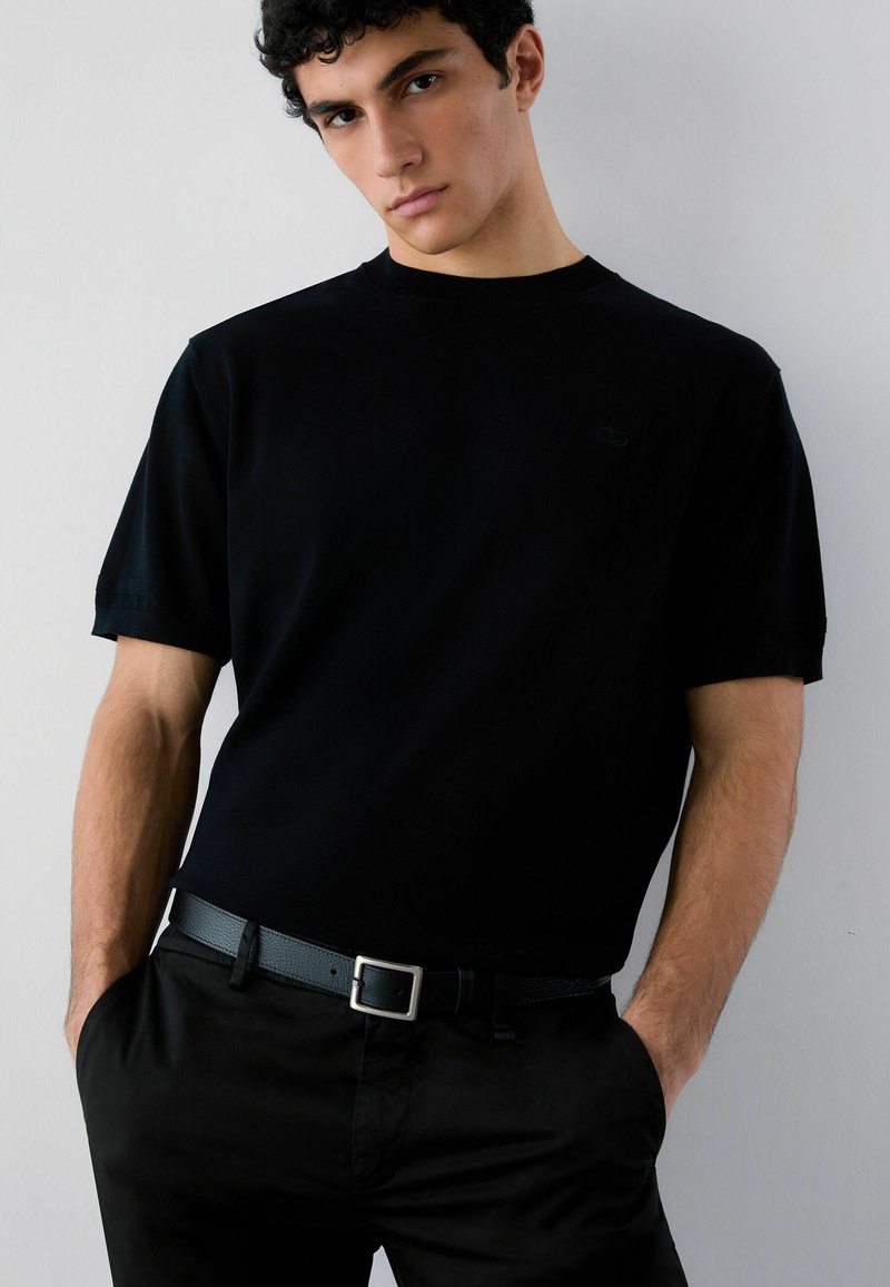 Junger Mann mit lockigem Haar, der ein schlichtes schwarzes Kurzarmshirt, schwarze Hose und schwarzen Gürtel trägt, steht mit den Händen in den Taschen.