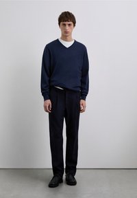 Marineblauwe gebreide trui met een V-hals, gedragen over een wit overhemd, gecombineerd met donkere broek en zwarte schoenen, staand tegen een effen achtergrond.