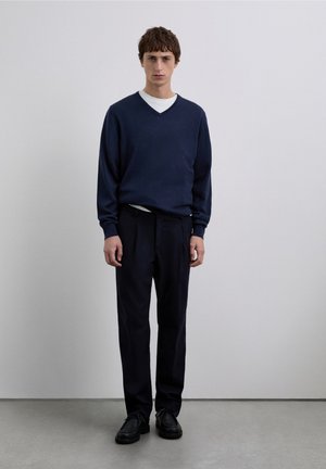 Pulover bleumarin tricotat cu guler în V, suprapus peste o cămașă albă, asortat cu pantaloni închiși la culoare și pantofi negri, stând în fața unui fundal simplu.